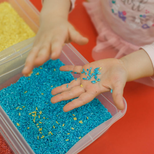 Toddler hands playing with rainbow rice in the sensory box. Baby's sensory educational kit