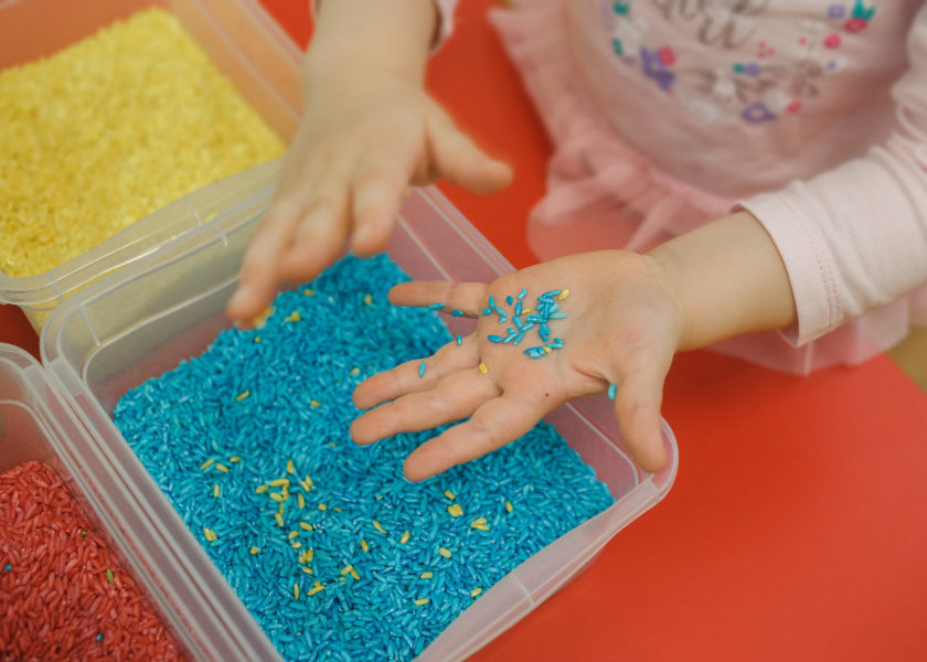 Child hands playing with colored rice in the sensory box. Baby's sensory educational kit