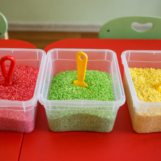 Sensory bin for toddlers with colourful rice on red table. Universal educational game in kindergarten.